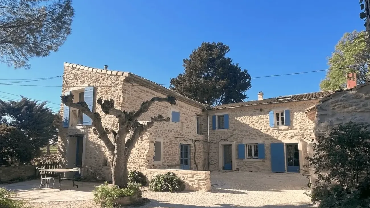 Cour et façade en pierre du mas aux volets bleus, sous le platane et les pins